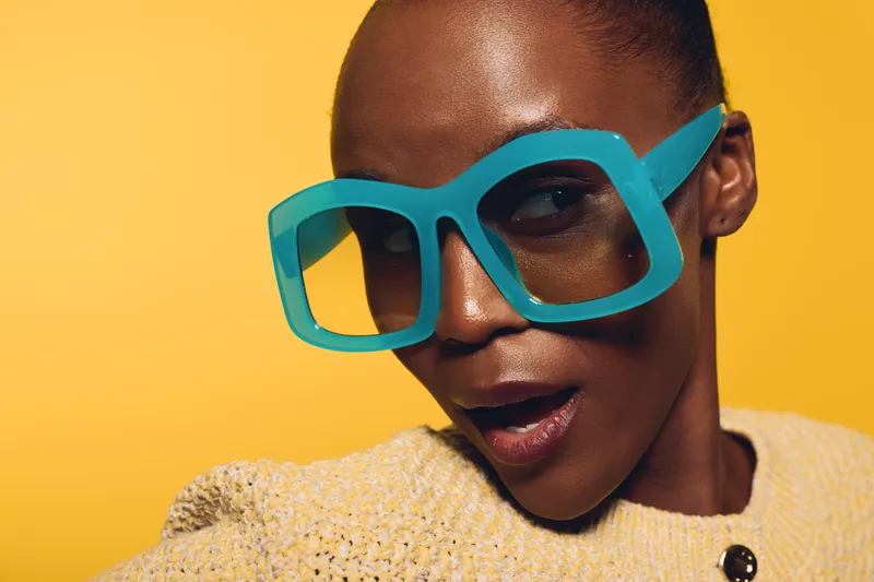 Close-up of a confident black woman with dark skin wearing oversized turquoise glasses and a beige textured sweater against a bright yellow background. Fashion and style concept.