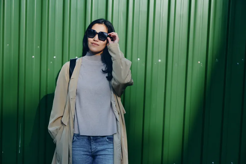 Woman wearing sunglasses and a beige coat stands against a green wall, casual fashion style in an outdoor scene, showcasing modern outfit details and confident demeanor