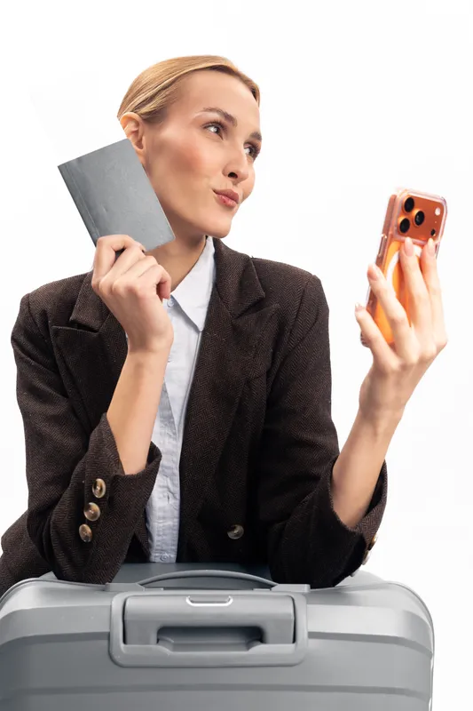 Young woman in brown jacket holds passport and smartphone, taking selfie while sitting on suitcase. Concept of travel preparation, vacation, and modern lifestyle with digital communication.