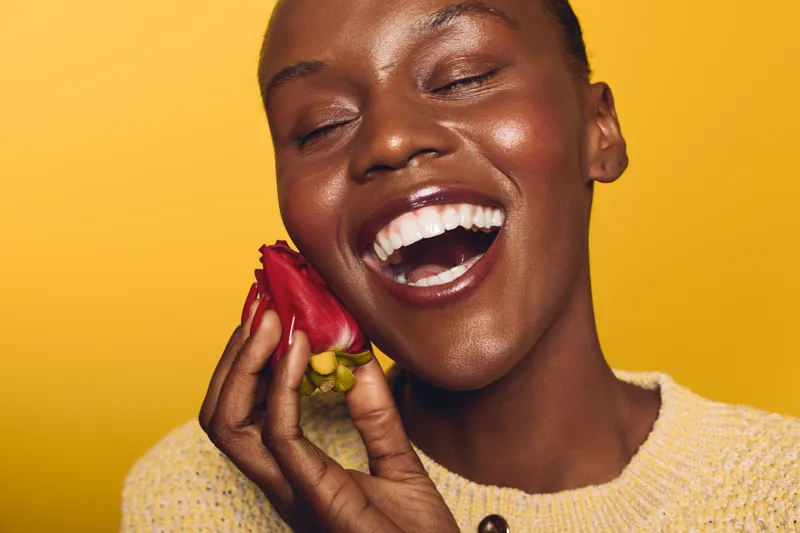Happy African woman with dark skin smiling with closed eyes holds red flower near face on yellow background. Concept of joy, beauty, and natural skincare with floral elements.