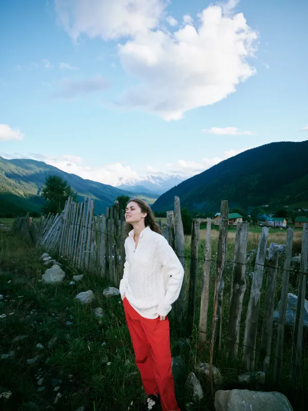 Woman wearing a white knit sweater and red pants stands beside a weathered fence in a rural valley with mountains under a bright blue sky, illustrating outdoor lifestyle, travel spirit, and serenity