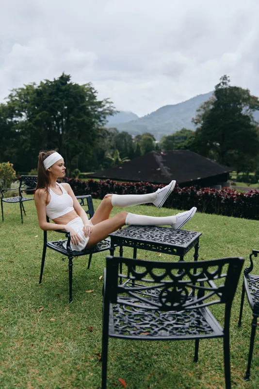 A young Asian woman in athletic attire sitting on a chair outdoors, exuding confidence and relaxation amidst a lush green backdrop, perfect for fitness and wellness themes