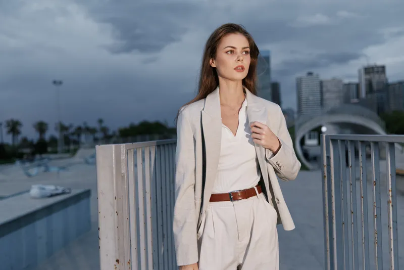 Confident businesswoman in tailored suit standing outdoors with urban background, expressing determination and focus during a cloudy day.