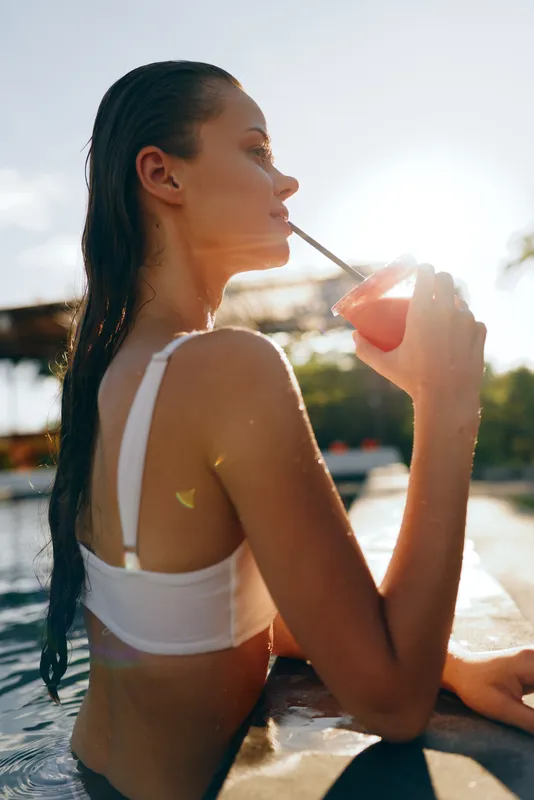 Young woman in white bikini top drinks juice with straw sitting by poolside enjoying sunny summer day with wet hair and relaxed lifestyle mood outdoors.