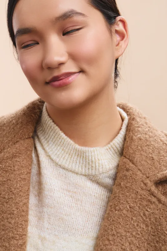 Asian woman wearing a cream turtleneck and beige coat smiles softly in a closeup studio shot, conveying warmth, calm confidence, and a serene, approachable mood ideal for modern lifestyle and fashion