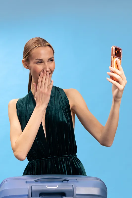 Young woman in dark green dress takes selfie with smartphone, smiling and covering mouth with hand. Blue background and suitcase visible. Concept of travel, happiness, and surprise.