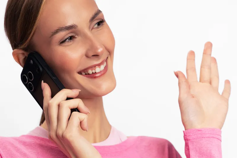 Smiling young woman in pink sweater talking on smartphone and waving hand isolated on white background. Concept of communication, happiness and friendly conversation.
