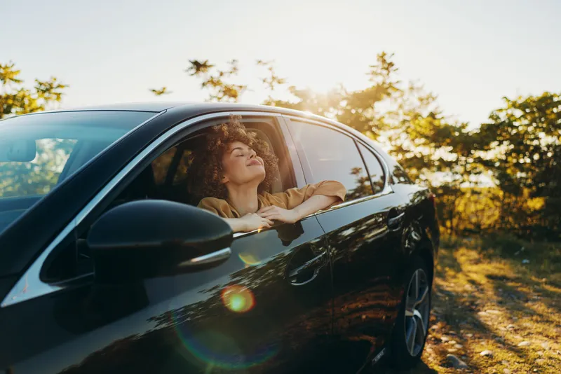 Woman leans from a car window in bright sunlight, retro film color vibe, casual mood, golden hour glow, outdoor lifestyle, serene moment isolated road, nature backdrop, warm tones