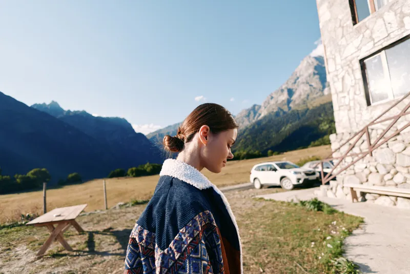 Woman standing near a stone chalet by a parked car in a grassy meadow, serene mountains in background. Outdoor portrait, travel lifestyle and rural countryside scene.