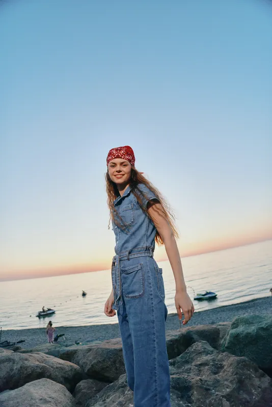Lifestyle woman in modern boho-western denim street style stands on rocky shore at sunset, captured with warm film color tones and casual confident expression.