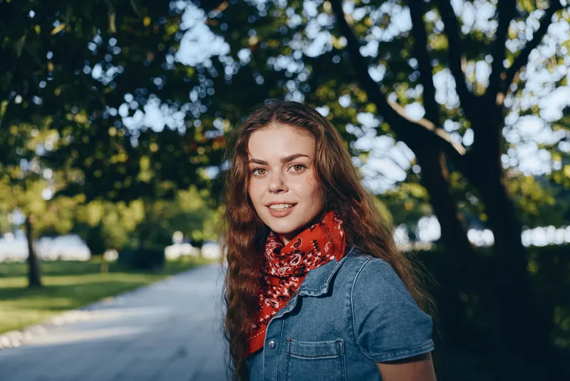 Lifestyle woman in modern boho-western denim street style wearing a red bandana outdoors with natural light and film color tones, smiling confidently in a sunny park setting.