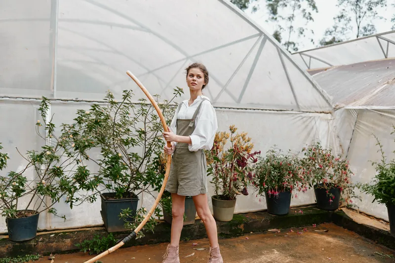 Confident young woman in a greenhouse holding a gardening tool, dressed in casual olive green overalls and a white blouse, showcasing a connection with nature
