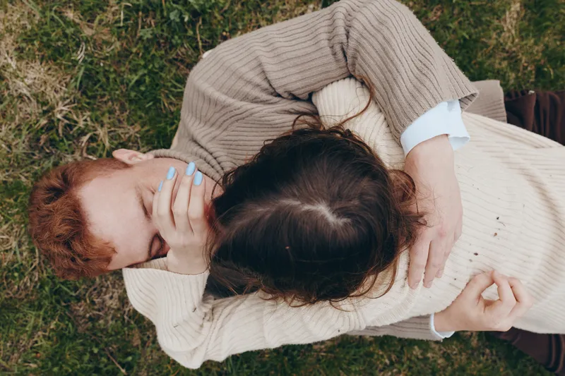 Two people share a warm hug on a grassy outdoor setting, casual sweaters and soft daylight convey comfort, connection, and everyday affection in a relaxed lifestyle moment