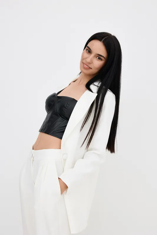 Smiling woman in white suit and black leather crop top posing confidently against plain background, with long straight hair and relaxed expression.
