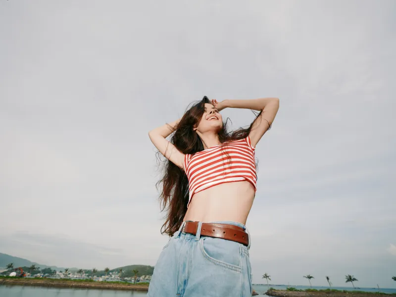 A young woman stands outdoors under a bright sky, wearing a striped crop top and denim jeans with a brown belt, arms raised, radiating carefree energy.