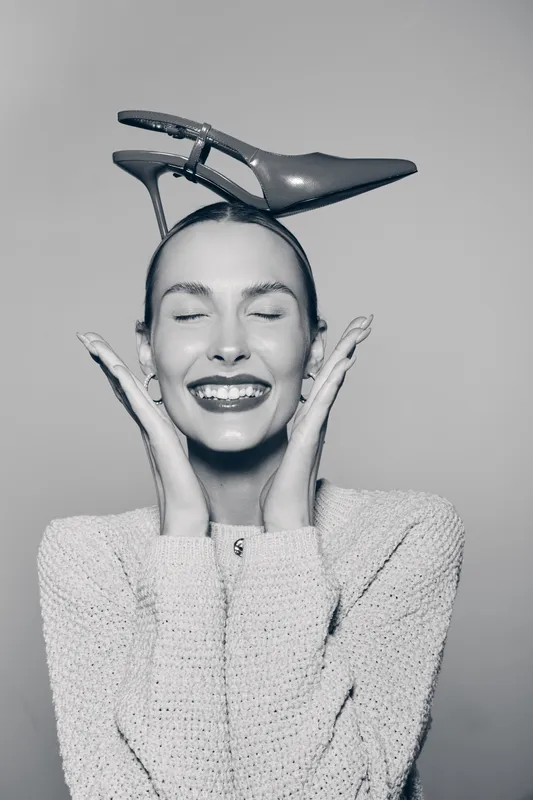 Happy white woman with light skin wearing knitted sweater smiles with closed eyes holding high heel shoe on head studio portrait. Concept of joy and fashion creativity.