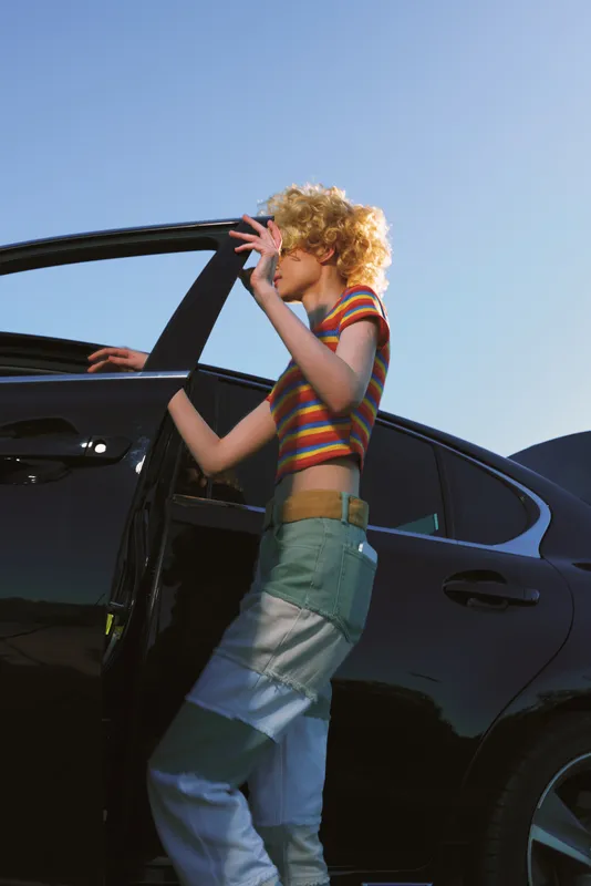 Woman with short curly blond hair steps from a sleek car wearing a striped crop top and vintage jeans, capturing a candid retro lifestyle moment under a clear blue sky today