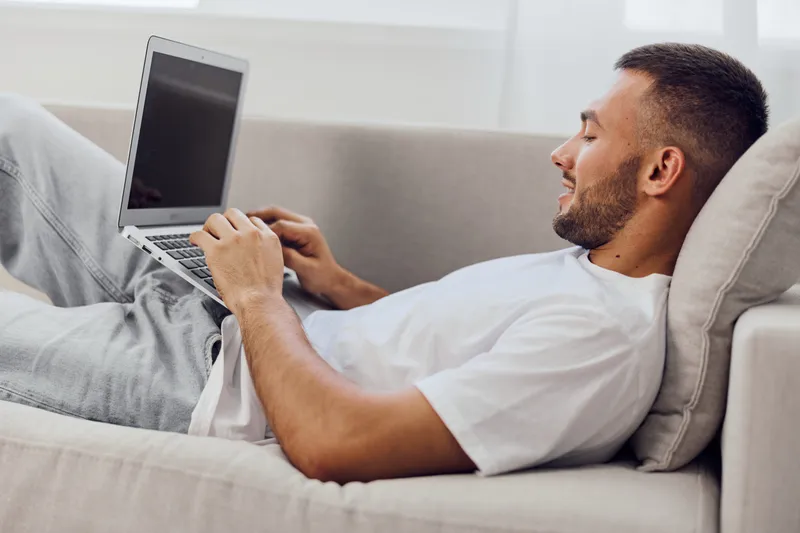 Relaxed man working on a laptop while sitting on a comfortable couch, wearing casual attire. A modern, minimalist setting enhances the cozy atmosphere and productivity.