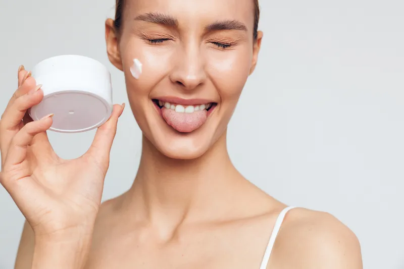 A young woman with clear skin holds a white cream container and applies moisturizer on her cheek while playfully sticking out her tongue against a plain light background.
