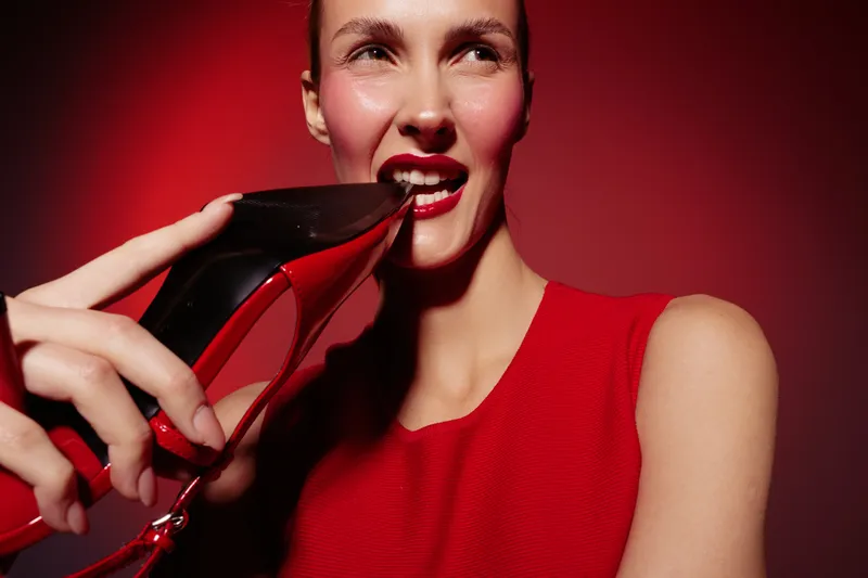 Young woman in red dress holds red high heel shoe near her mouth against red background. Fashion concept with bold makeup and confident expression in studio lighting.