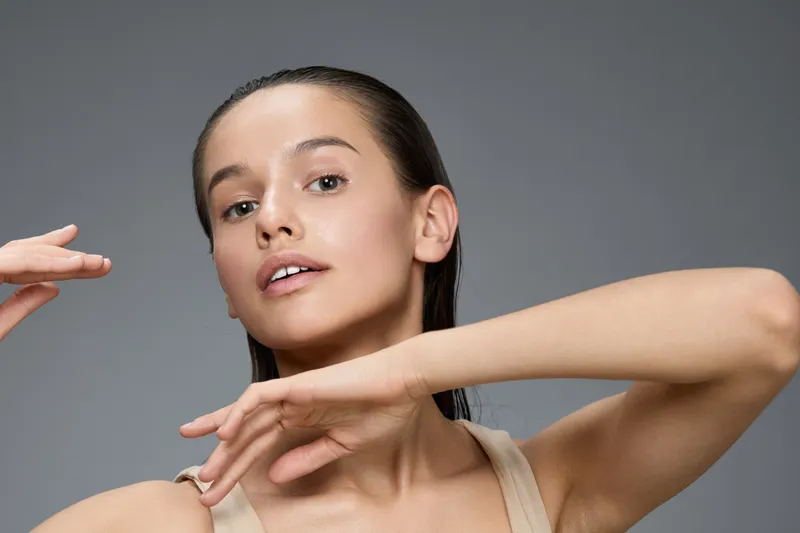 Young woman with glowing skin posing gracefully against a neutral background, showcasing beauty, confidence, and elegance in a minimalistic style