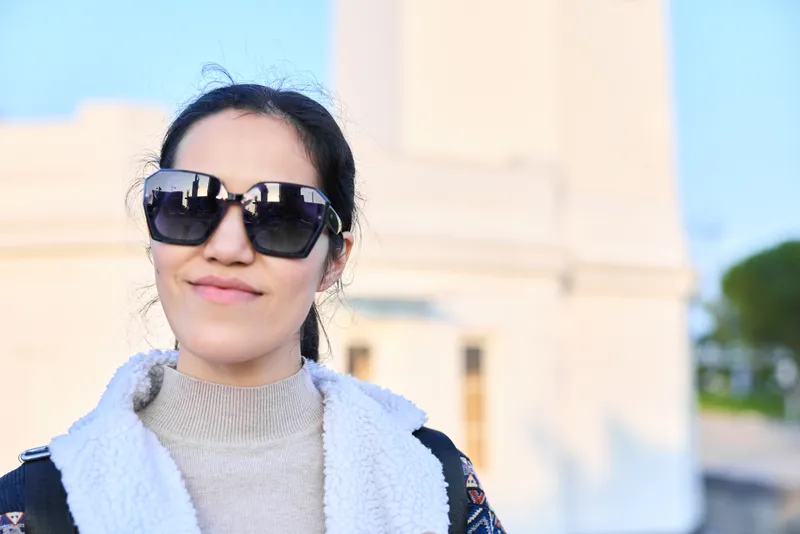Sunglasses wearing woman outdoors with knit sweater and scarf near a light monument, showcasing casual fashion and a relaxed sunny daytime mood in an urban setting