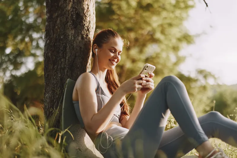 Young woman enjoying her smartphone in nature, wearing casual athletic wear, smiling, sitting against a tree in the warm sunlight