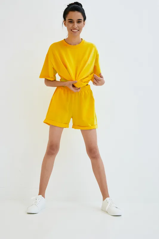 Young woman wearing bright yellow casual outfit stands playfully against white background, smiling and tying her shirt in a relaxed pose with white sneakers.