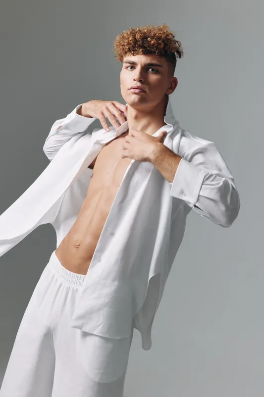 Young man with curly hair wearing open white shirt and white pants posing confidently. Studio portrait with plain gray background showing fashion style and modern grooming of male model.