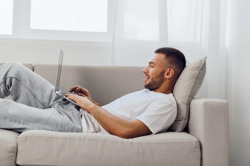 Relaxed man lying on a sofa, working on a laptop while enjoying a cozy and modern indoor environment with natural light pouring in from the windows.