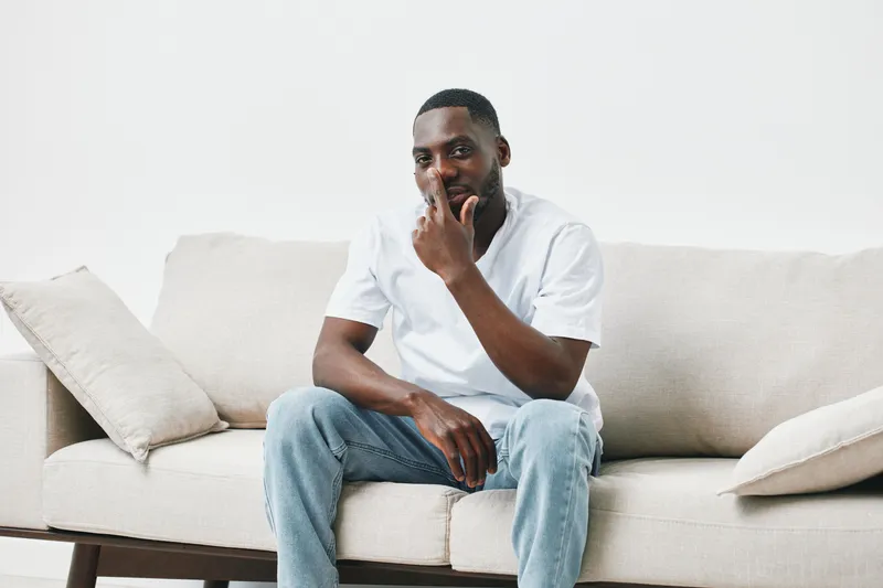 A thoughtful Black man sitting on a cozy sofa, wearing a casual white shirt and jeans, reflecting a sense of calm and introspection in a modern interior