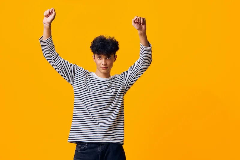 young man celebrating with raised arms, wearing striped shirt, curly hair, cheerful expression, bright yellow background, casual style
