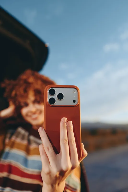 Woman lifestyle, hands holding smartphone with red case outdoors, close view of fingers forming a peace sign, blurred woman in plaid jacket behind, bright sky and travel mood.