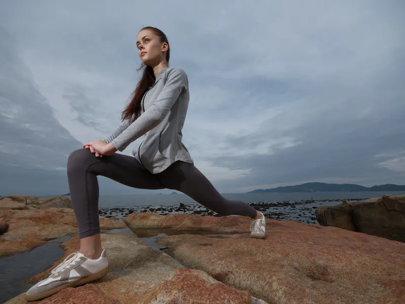 Young female athlete stretching on rocks by the sea, dressed in sportswear, showcasing determination and focus, with a cloudy background