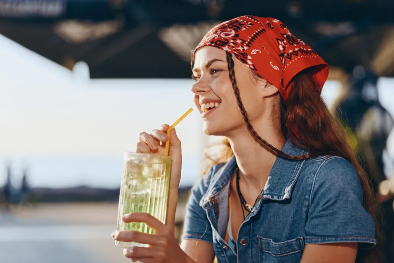 Lifestyle woman in modern boho-western denim street style enjoying a refreshing drink with a bright smile and film color warmth outdoors in casual summer setting.