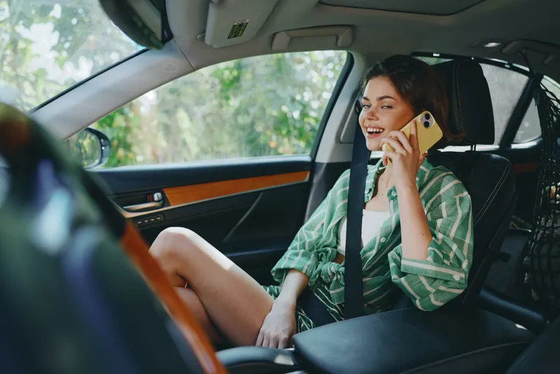 Young woman sits in the front passenger seat, smiling and talking on a smartphone during a sunny drive.