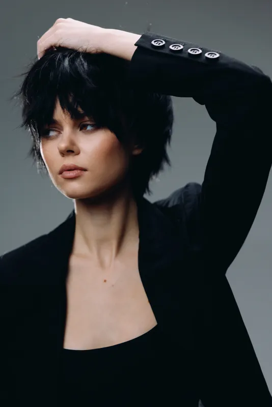 Young woman with short black hair in black jacket posing with raised arm in studio, stylish and confident mood