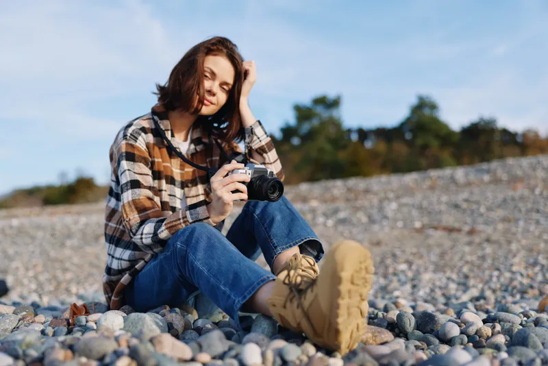 Photographer woman outdoors on a pebble beach, holding a camera, casual plaid shirt and boots, capturing a relaxed outdoor portrait amid nature today.