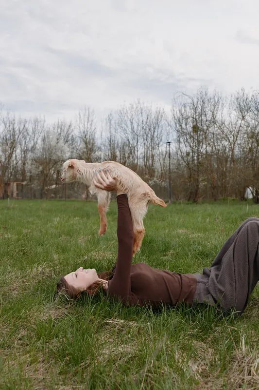 Relaxed outdoor lifestyle scene shows a person lying on grass in a meadow, lifting a goat kid with joyful expression, natural light, serene countryside background