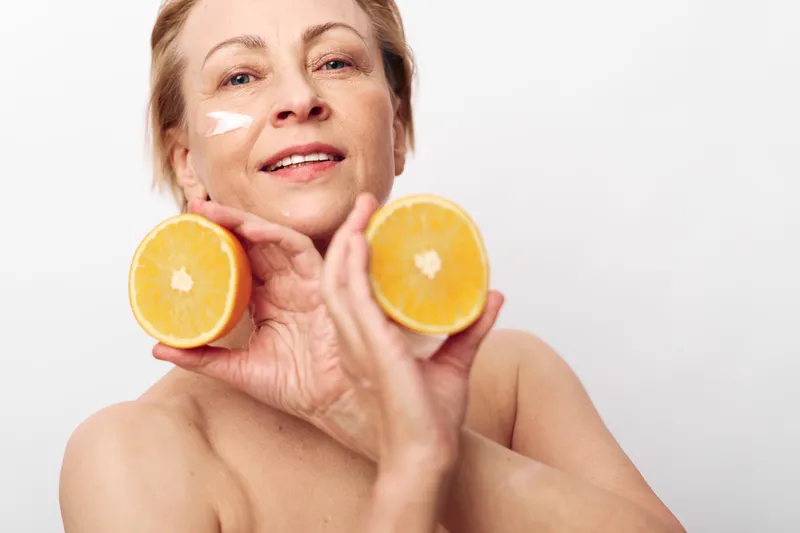 Smiling mature woman with natural skin holds two orange halves near her face with white cream on cheek, isolated on white background. Concept of skincare, health, and natural beauty care.
