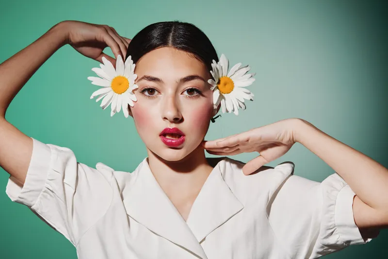 Stylish young woman with dark hair and bold red lipstick poses with daisies by ears. Green background highlights fresh beauty close-up. Concept of fashion and creative self-expression.