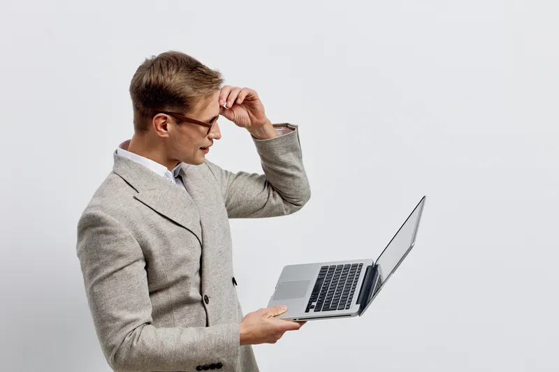 Professional young man wearing glasses and gray blazer looking at laptop screen in surprise or confusion isolated on plain background studio portrait. Business, technology, work lifestyle concept