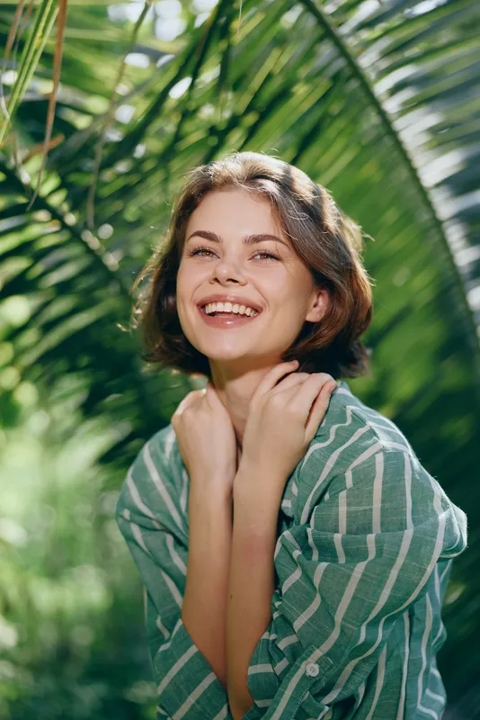 Joyful portrait of a smiling woman outdoors, sunlight on her face, framed by lush tropical leaves.
