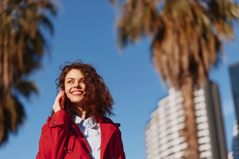 Young woman with curly hair smiles happily while standing outdoors in a red coat under palm trees on a sunny day.