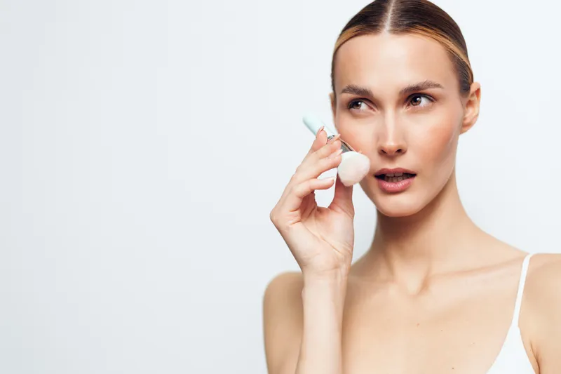 Young woman with clean skin applies white facial brush to her cheek looking sideways isolated on white background beauty skincare concept.