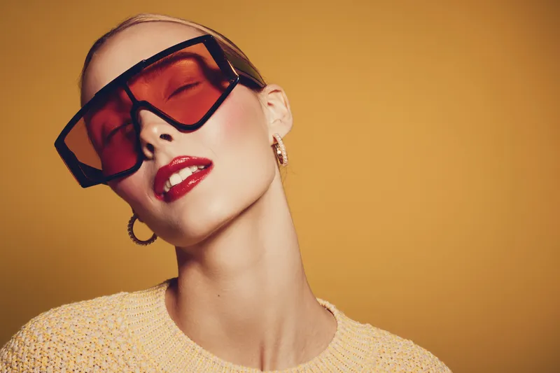 Stylish woman with shaved head wears oversized red sunglasses and yellow sweater. She poses confidently against a warm yellow background. Concept of fashion, beauty, and modern style.