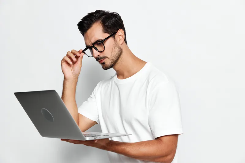 Thoughtful young man wearing glasses looking at a laptop while adjusting his glasses, isolated on plain solid white background. Technology and lifestyle concept.