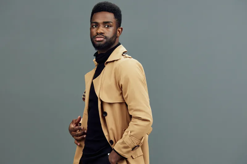 Stylish young man in a beige trench coat against a neutral background, embodying confidence and modern fashion trends during the fall season