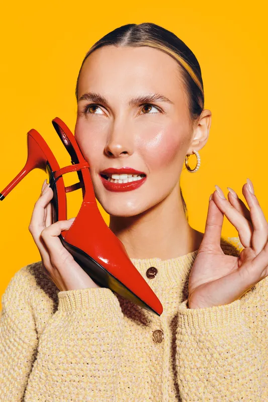 Young woman with slicked hair holds red high heel shoe near face, wearing beige sweater and gold hoop earrings. Bright yellow background creates vibrant fashion and style concept.