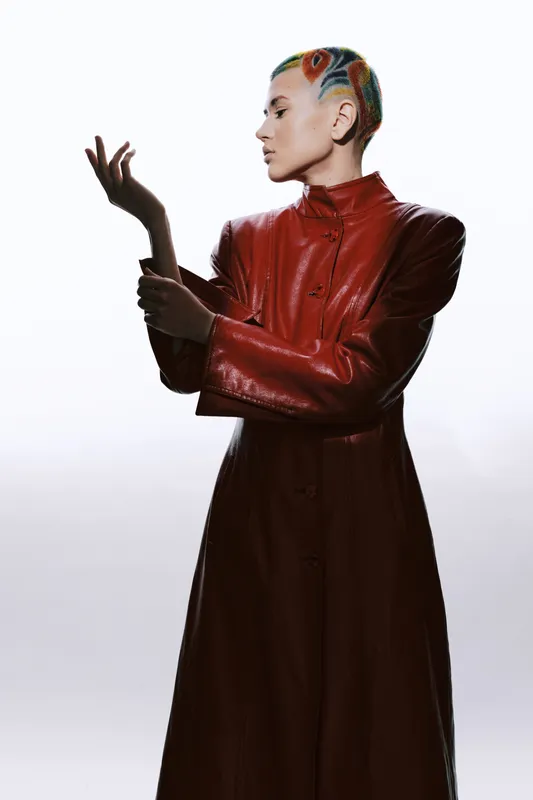 Artistic woman with colorful braided hair wearing a long red leather coat, posing thoughtfully against a bright white background in a modern studio setting.